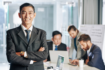 Portrait of smiling confident Asian entrepreneur standing with arms crossed, his colleagues working on business development strategy in background