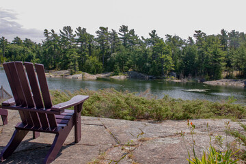 Fototapeta premium Two black Muskoka chairs sitting on a wood dock facing a lake. Across the calm water is a white cottage nestled among green trees. Canada flag is visible