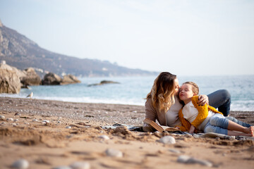 Woman read the book and relax in the blanket in the beach against stone while kid listen. Mother hugging with baby. Happy leisure to relax with family on the sea shore