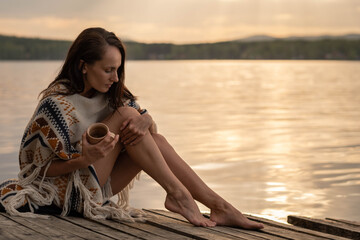 A beautiful dark-haired woman sits on an old wooden pier in the early hours of dawn holding a ceramic mug of fragrant tea, stroking her leg