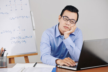 Bored or tired young school teacher preparing for online class and checking documents on laptop