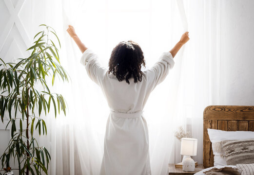 Perfect Morning. Unrecognizable woman in white bathrobe opening window curtains at home - Powered by Adobe