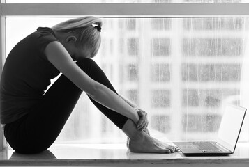 Sad depressed young woman with her head down sitting alone with a laptop computer by the window in...