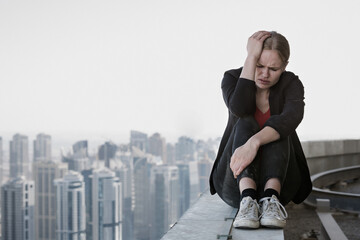 Sad young businesswoman sitting alone on edge of skyscraper rooftop with hand on the face against modern city high-rise view background. Work stress and failure. © PKpix