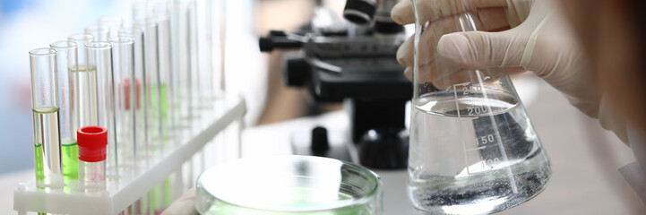 Chemical experience in laboratory, water sample. Chemist holds in his hands flask with clear liquid, next to it are tubes with liquids. In background is equipment for conducting water analysis.