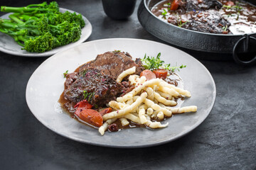 Traditional German braised beef cheeks in brown red wine sauce with noodles and broccoli offered as closeup on a modern design plate and stewpot on an old rustic board