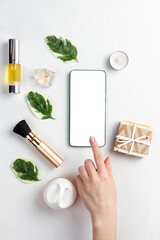 Fototapeta na wymiar Cosmetics and modern technologies. A woman's hand taps the screen of a smartphone, which is lying on a white background with cosmetics and leaves. The concept of organic cosmetics