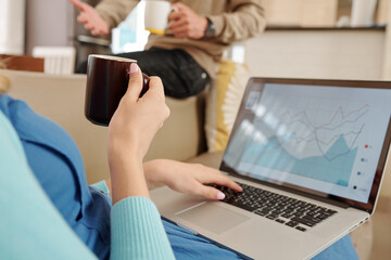 Woman sitting on sofa, analyzing financial chart on laptop screen, drinking coffee and talking to...