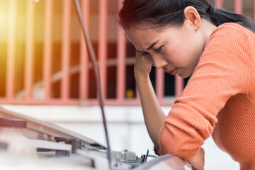 Stressed asian woman while looking at break down car engine. Side portrait of young woman looking under the hood of car engine defective on the road with sunlight.