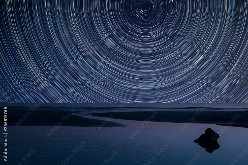 Wall mural digital composite image of star trails around polaris with vibrant landscape of low tide beach