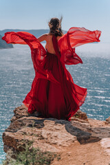 A girl with loose hair in a red dress waved her skirt on the yellow rocks overlooking the sea. Sunny path on the sea from the rising sun.