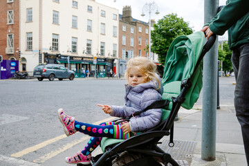 Cute little toddler girl sitting in baby stroller while walking with dad on the streets of big...