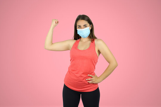 Young beautiful woman standing over pink isolated background wearing sport chlotes and mask. Doing a gesture with his arm of strength and victory