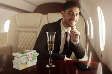 Handsome billionaire in suit and tie sitting next to window in his private jet with glass of champagne and packs of banknotes, smiling happily, satisfied with income