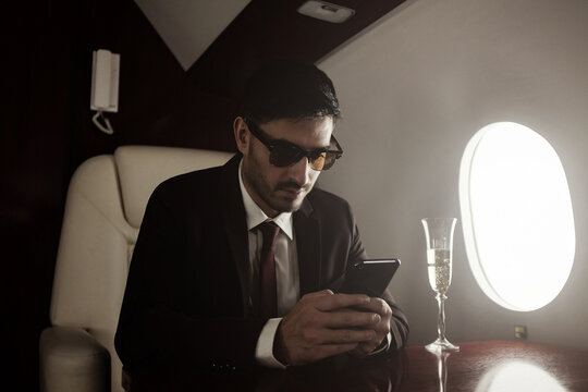 Rich businessman sitting on board of private jet in armchair at table, drinking champagne and reading messages or checking business news on smartphone