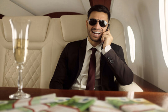 Young happy billionaire on board of his private jet sitting at table with champagne and euros in cash, talking on phone and laughing happily, enjoying financial success