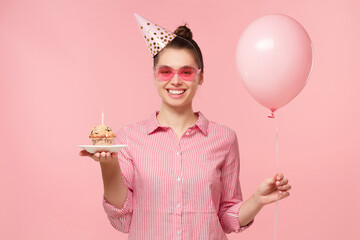 Young happy female in striped shirt, wearing colored glasses and birthday hat, standing with...
