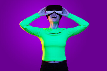 Obraz premium Young excited girl wearing green neon top and virtual reality headset smiling and laughing with joy, looking at screen , isolated on purple background