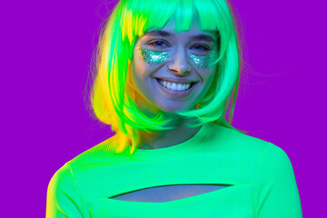 Headshot portrait of young teenage girl, dressed in neon green top, yellow hair and glitter makeup on cheeks, having fun, isolated on purple background
