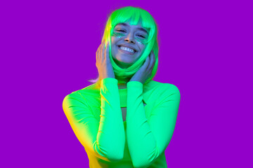 Close-up portrait of young happy teenage girl wearing neon green top and yellow wig, smiling and touching her hair as if feeling shy, isolated on purple background