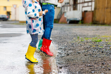 Close-up of three children, toddler girl and two kids boys wearing red, yellow and green rain boots and walking during sleet. Happy siblings jumping into puddle. Having fun outdoors, active family