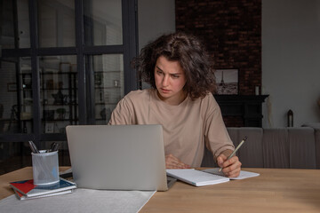 Young student girl with dark curly hair sitting at the table with laptop thoughtfully studying at cozy home. Online learning concept