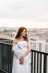 Beautiful girl on the terrace in a white blanket at sunset. Girl posing background fashion. Mood girl wrapping with blanket on balcony. Terrace mood.