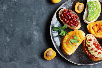 Sweet toast with jam and various fruits apricots,bananas,pomegranates and avocado on the dark background copy space top view.