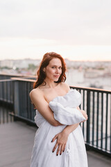 Beautiful girl on the terrace in a white blanket at sunset. Girl posing background fashion. Mood girl wrapping with blanket on balcony. Terrace mood.