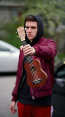 Obraz premium Young unshaven man in a red jacket with a black hood with a ukulele in his hand