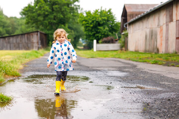 Little toddler girl wearing yellow rain boots, running and walking during sleet on rainy cloudy day. Cute happy child in colorful clothes jumping into puddle, splashing with water, outdoor activity