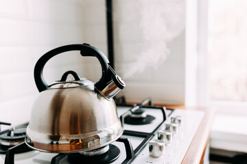 The kettle boils, steam from metal teapot on gas oven. Bright kitchen interior. White modern dining room. Wooden complete kitchen with gas oven.
