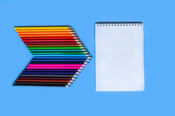 Stationery. The pencils. A pointer consisting of colored pencils pointing towards a notepad in a cage. Stationery template on a blue background.