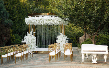 Luxurious yellow-gold wedding arch decorated with white flowers in nature among the trees. wedding ceremony. white piano on the nature. chairs for guests on the street.
