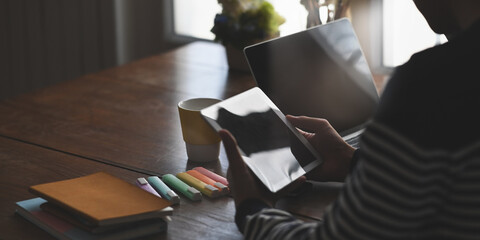 A young man is holding a computer tablet while sitting in front of his computer laptop at the wooden working desk.