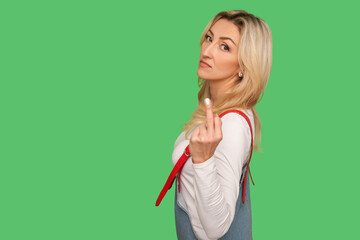 Portrait of impolite rude adult woman in denim overalls rejecting communication, showing middle finger to express disrespect and hate, vulgar gesture. indoor studio shot isolated on green background
