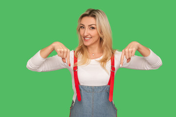 Look, advertise below! Portrait of positive energetic adult woman in stylish denim overalls pointing down, showing place for promotional message. indoor studio shot isolated on green background
