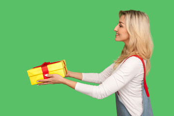 Take this present! Side view of generous beautiful adult woman with friendly smile giving wrapped box, congratulating and offering gift, delivering goods. studio shot isolated on green background