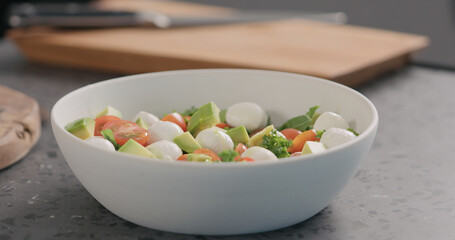 man add oil into salad with kale, avocado, mozzarella and tomatoes in white bowl on kitchen countertop