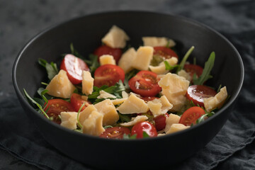 Fresh salad with cherry tomatoes, arugula and hard vintage cheese in black bowl on linen napkin