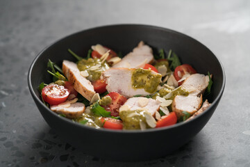 Salad with chicken, tomatoes and arugula in black bowl on concrete background