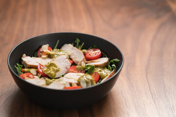 Salad with chicken, tomatoes and arugula in black bowl on walnut wood background with copy space