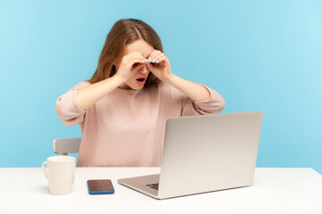 Curious woman employee looking at laptop screen through binoculars hand gesture, viewing interesting content, searching and discovering on internet. indoor studio shot isolated on blue background