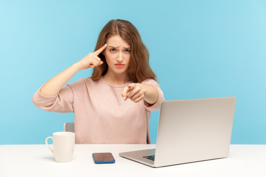 You Are Idiot! Woman With Sarcastic Look Sitting At Workplace And Making Stupid Gesture, Pointing To Camera, Teasing And Accusing Senseless Dumb Mind. Indoor Studio Shot Isolated On Blue Background