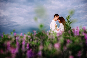 Loving couple on a background of blue sky with clouds. A guy and a girl are surrounded by purple flowers. Lifestyle concept. Honeymoon of lovers. Rest at nature