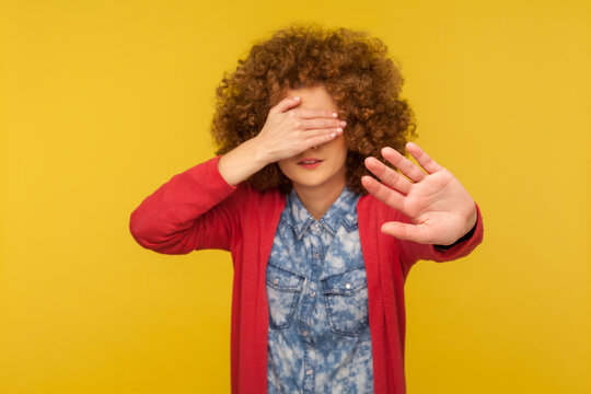 No, don't want to look. Portrait of scared woman with curly hair covering eyes and showing stop gesture, feeling shy afraid to watch, ignoring troubles. studio shot isolated on yellow background