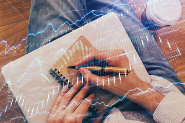 Double exposure of hands writing in notepad with stock market chart.