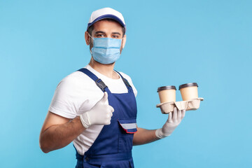 Fototapeta na wymiar Delivery service. Side view, friendly courier in overalls and mask holding coffee, wearing safety gloves offering drinks in disposable cups and showing thumbs up, like gesture. indoor shot, isolated