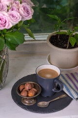 A bouquet of roses in a vase stands with a cup of tea with sweets on the windowsill of the house - a sweet home