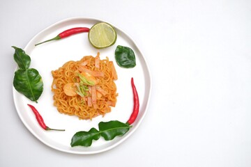 Top view a spicy dried instant noodle in a white plate  with fresh kaffir lime leaves,red hot chilli and a half cut of a lemon,white background with copy space